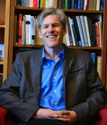 A smiling man sitting in a red chair in front of a full bookcase.