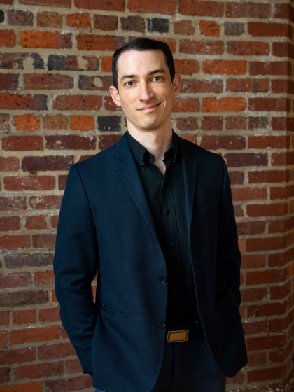 Ben Sarbey standing in front of a brick wall with a black shirt and navy blazer. 